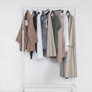 A white clothing rack holds a selection of neutral and earth-toned garments such as coats, shirts, dresses, and pants, all hanging on black hangers in front of a plain white background.