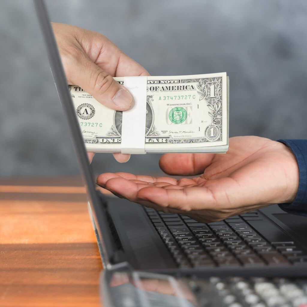 A hand passes dollar bills through a laptop screen to another open hand, representing digital payment or online money transfer.