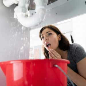 A concerned woman watches water leaking from pipes beneath a sink into a red bucket, indicating a plumbing issue.