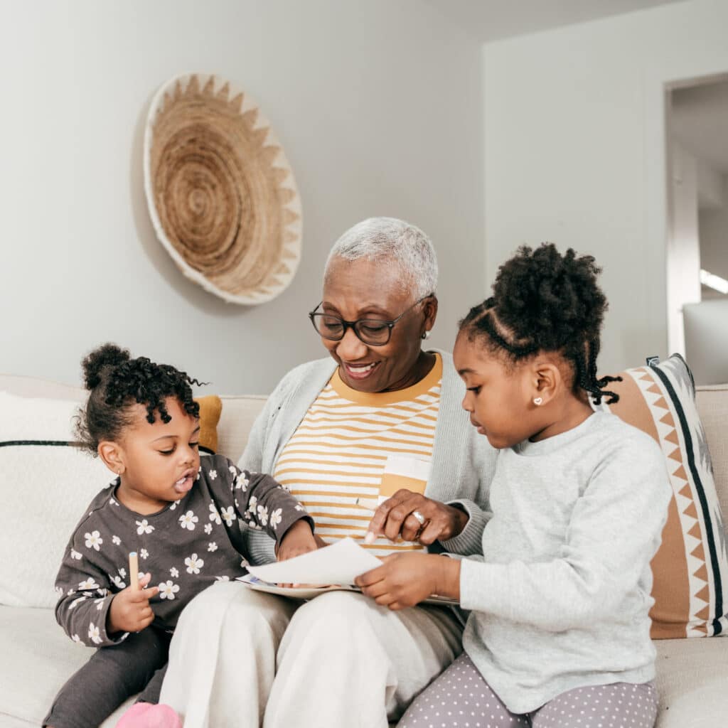 An older woman with short gray hair and two young girls smile together on a couch, sharing a book in a cozy living room.