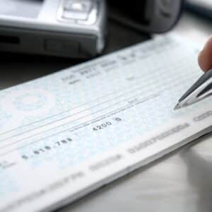 Someone fills out a blank check with a pen. The check and pen are in sharp focus, while the background objects appear blurred.