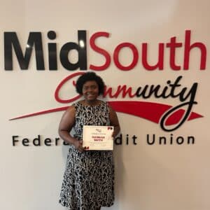 A woman in a black and white dress holds a certificate in front of a wall with MidSouth Community Federal Credit Union in large letters.