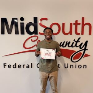 A person stands smiling in front of a MidSouth Community Federal Credit Union sign, holding a certificate with the name Zachary Johnson on it.