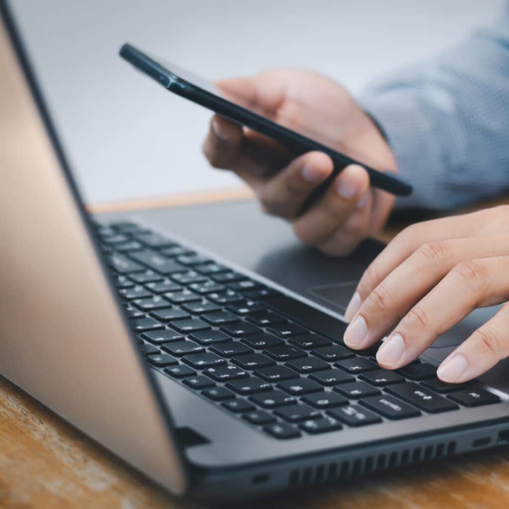 Someone in a long-sleeve shirt works on a laptop, typing with one hand while holding a smartphone in the other; both devices rest on a wooden table.