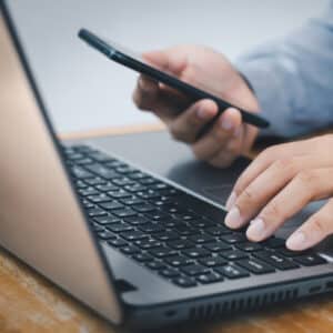 Someone in a long-sleeve shirt works on a laptop, typing with one hand while holding a smartphone in the other; both devices rest on a wooden table.