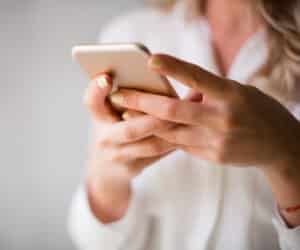 A person with light-colored nail polish holds a smartphone in both hands, typing with their thumbs. The person is wearing a white shirt, and the background is blurred.